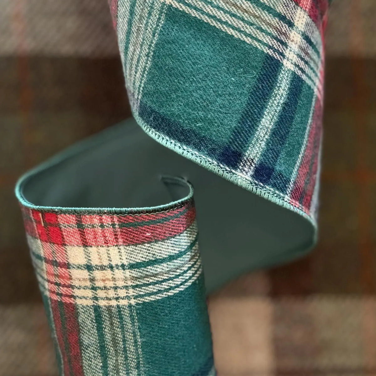 A close-up of a folded plaid fabric or ribbon with a tartan pattern in teal green, red, cream, and navy, shown against a blurred Harris Tweed plaid background.