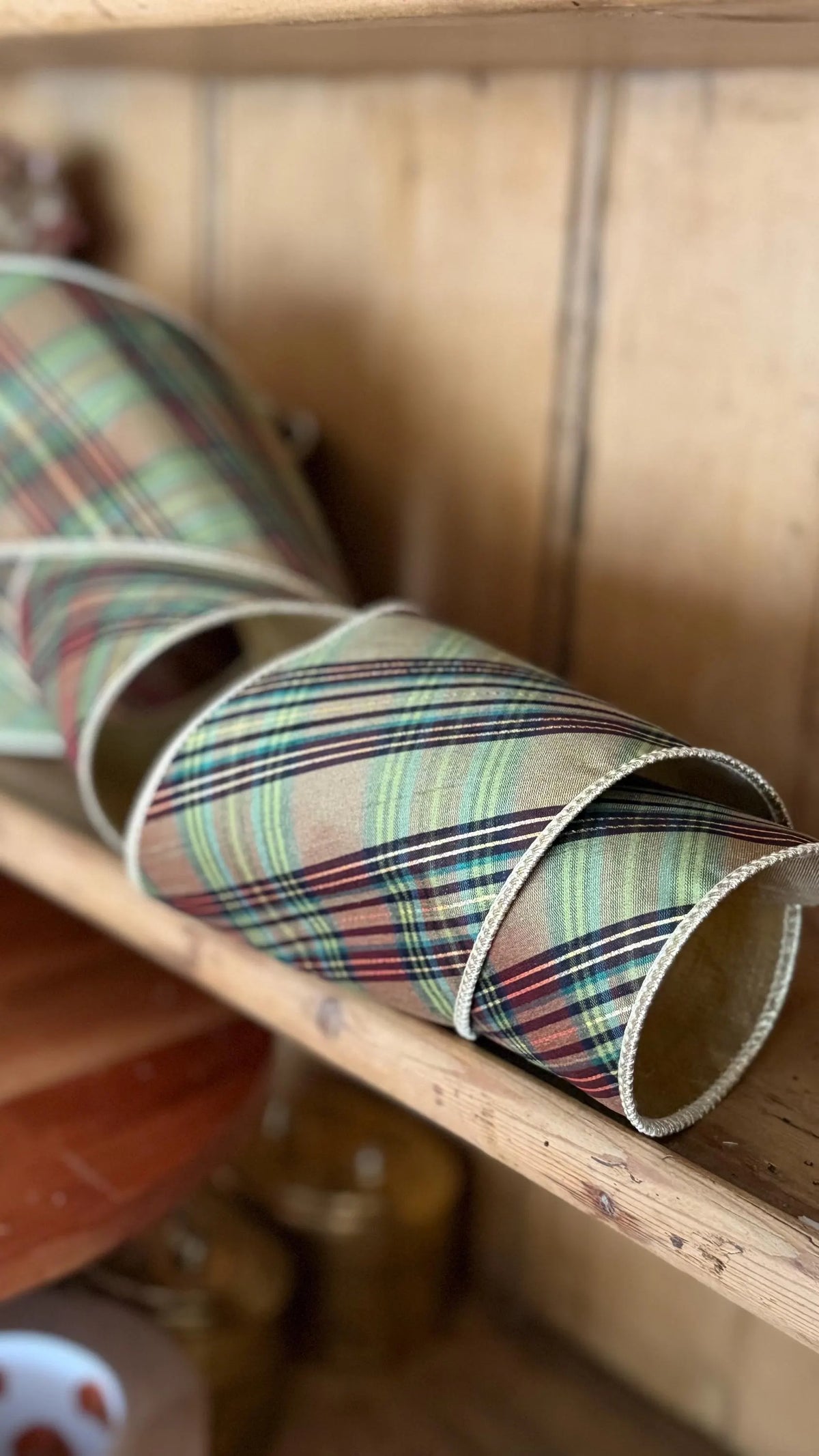 A close-up of a rolled piece of plaid fabric or ribbon with a tartan pattern in green, red, blue, and beige, resting on a wooden shelf.
