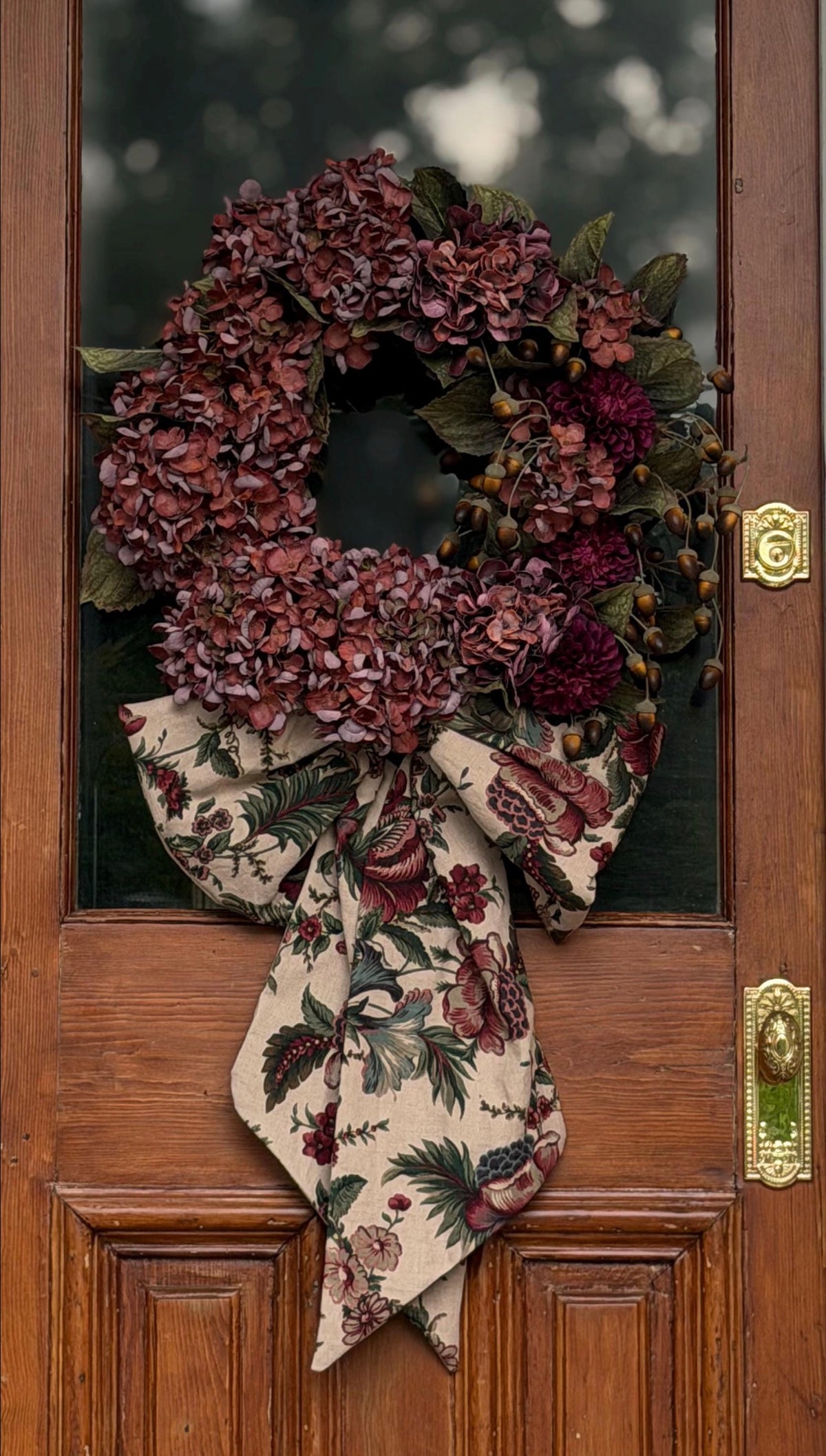 A decorative autumn wreath hanging on a wooden front door. The wreath features clusters of dark burgundy hydrangea-like flowers, green leaves, small brown acorn details, and a large fabric bow at the bottom with a beige floral print.