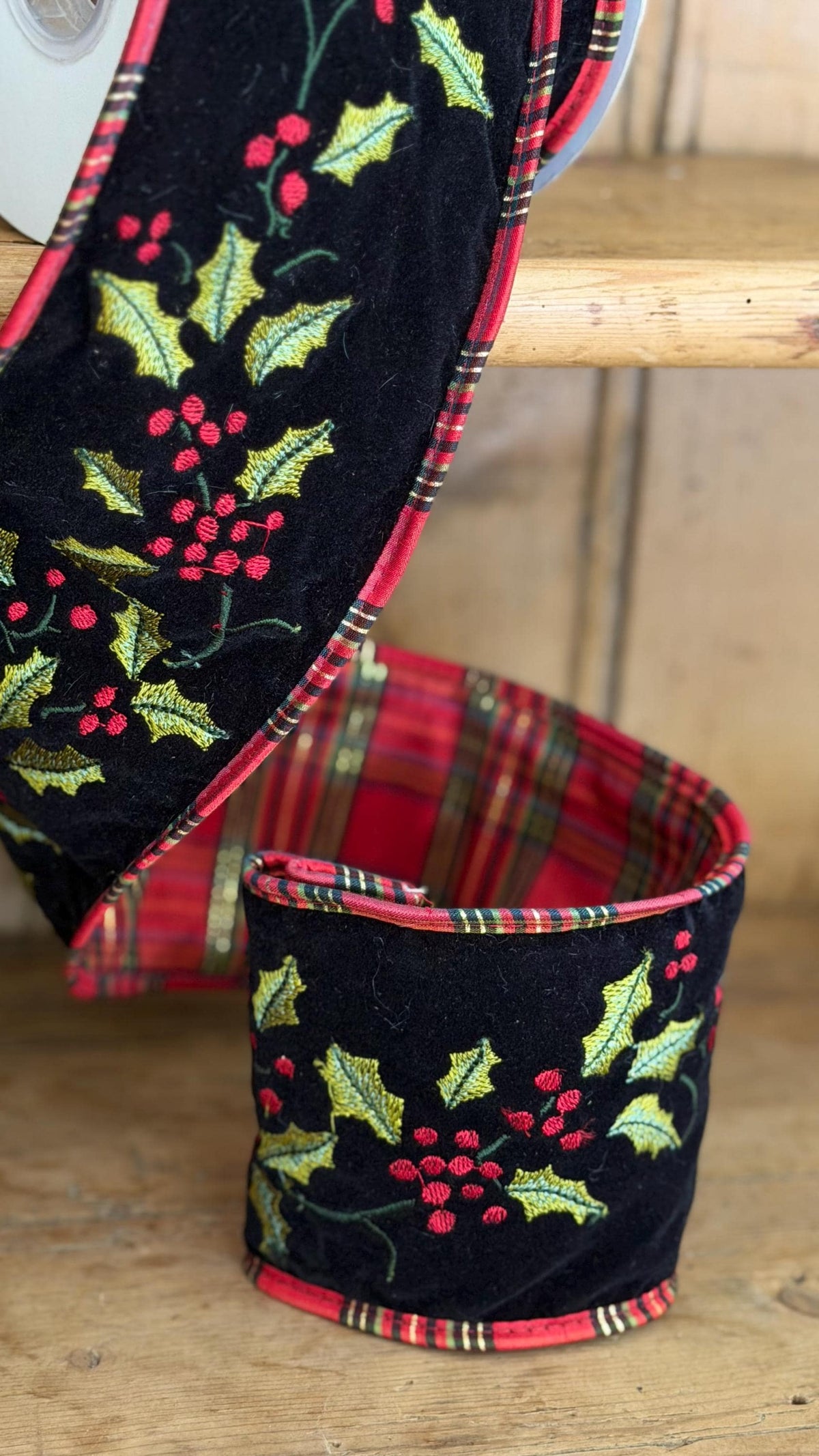 A close-up of a black velvet holiday garland and matching cup holder set with embroidered holly leaves and red berries, featuring red and green plaid trim, displayed on a rustic wooden shelf.