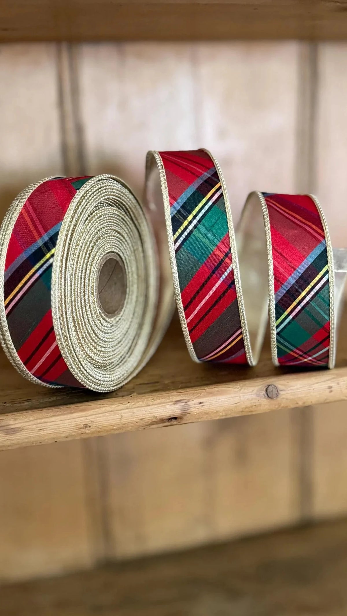Three rolls of plaid ribbon in red, green, black, and gold tartan patterns, displayed on a rustic wooden shelf.