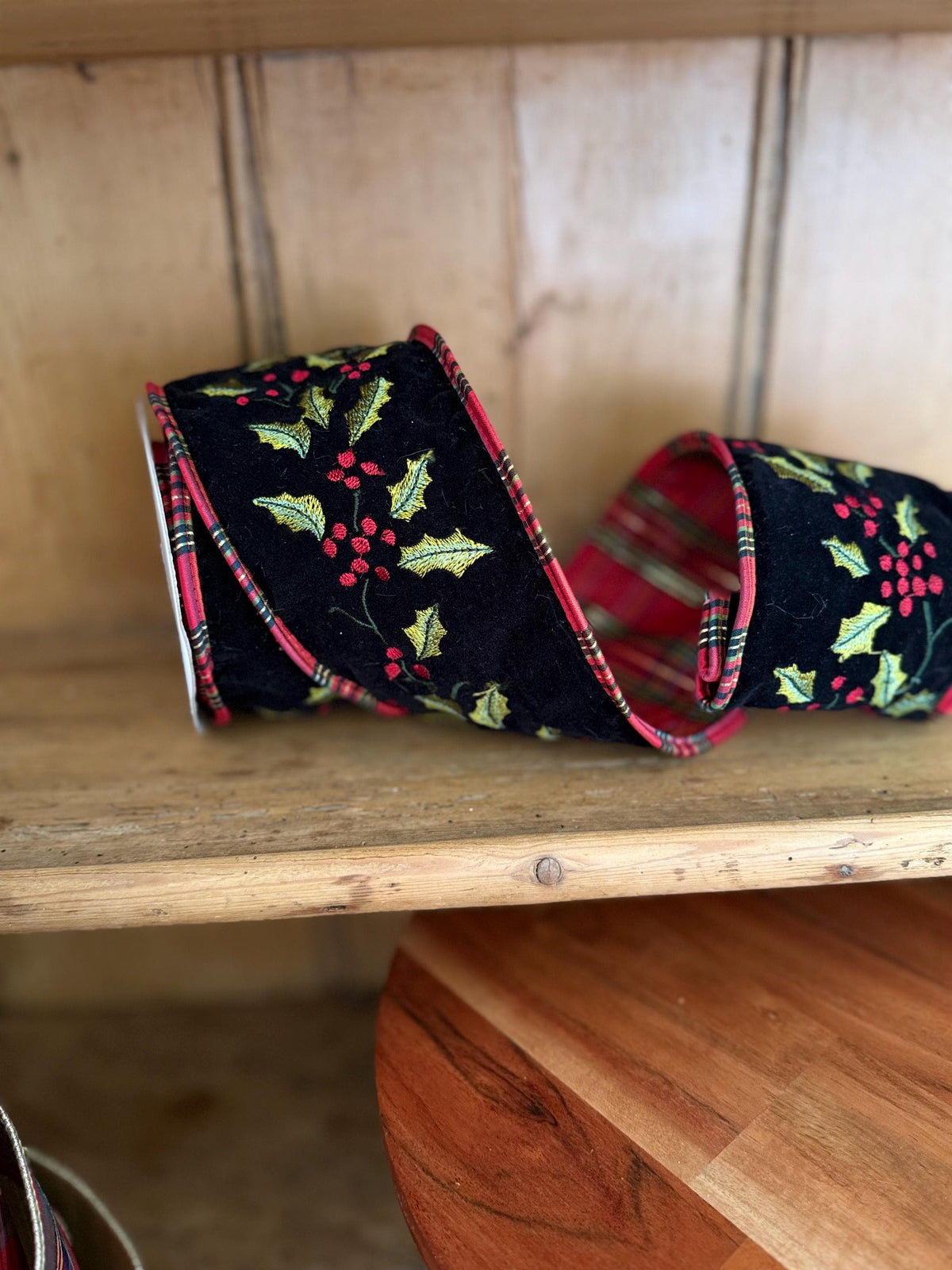 A festive Christmas-themed ribbon with a black velvet background, green holly leaves, red berries, and red tartan plaid edges, displayed on a wooden shelf.