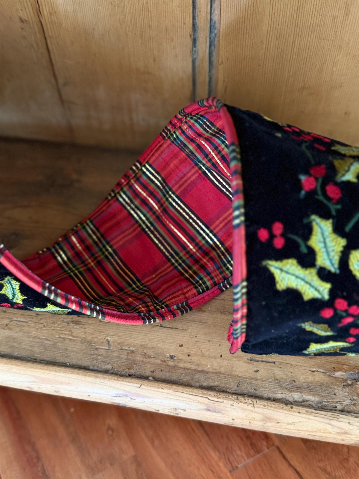 A close-up of red tartan ribbon reverse, showing two different holiday-themed patterns: a red and green tartan plaid side and a black velvet side with a green holly and red berry print. The 4inch black velvet ribbon is resting on a rustic antique wooden pine hutch from Wales.