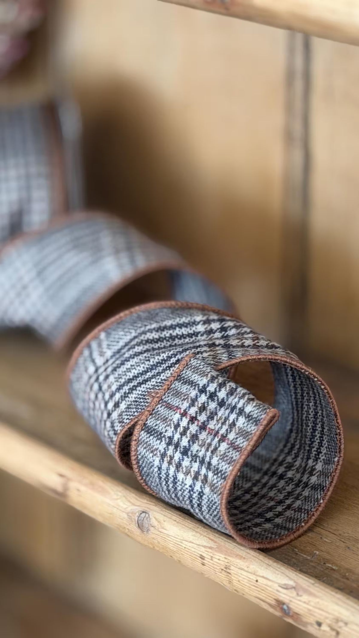 A close-up of a rolled woven strap or belt made from gray, black, and white plaid fabric with brown trim, resting on antique wooden surface.