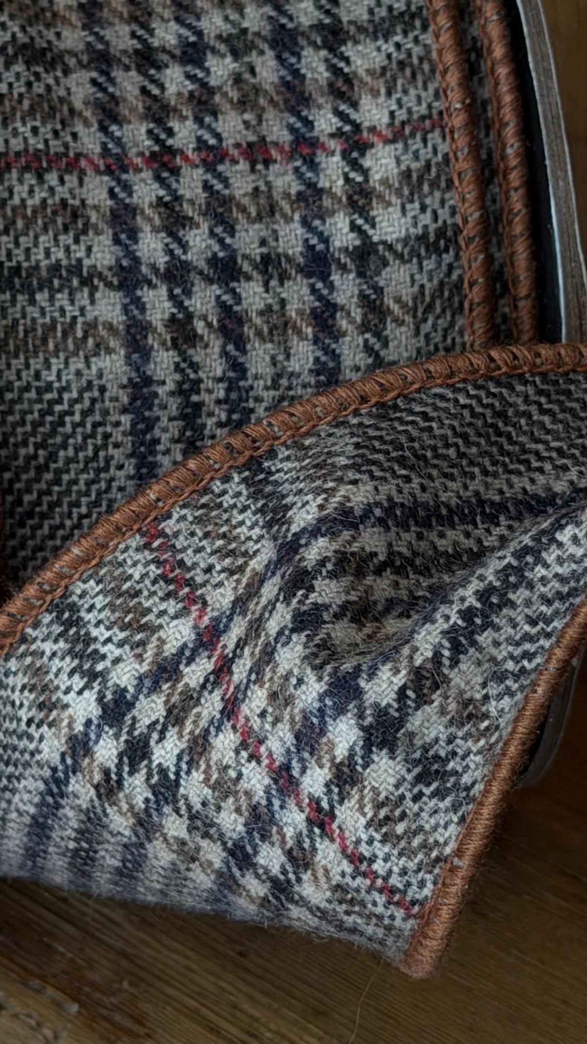 Close-up of a textured tweed fabric with a brown, beige, and gray houndstooth pattern, featuring a brown braided trim and a thin red accent stitch, laid on a wooden surface.
