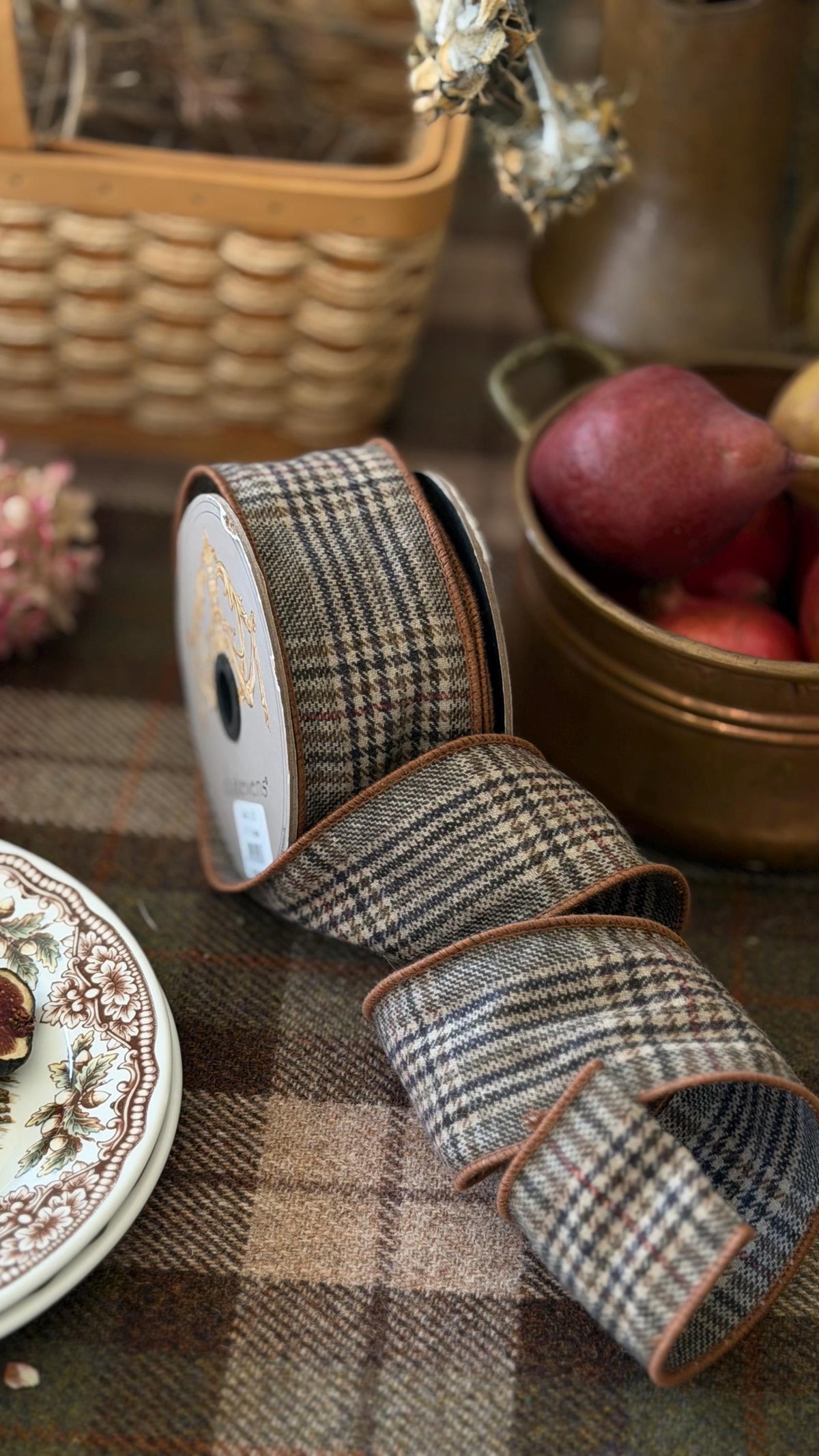 A roll of plaid ribbon with brown trim, unrolled slightly on a matching plaid surface. The ribbon has a beige, brown, and gray check pattern. Pears in copper pot, basket with winter twigs, china with figs.