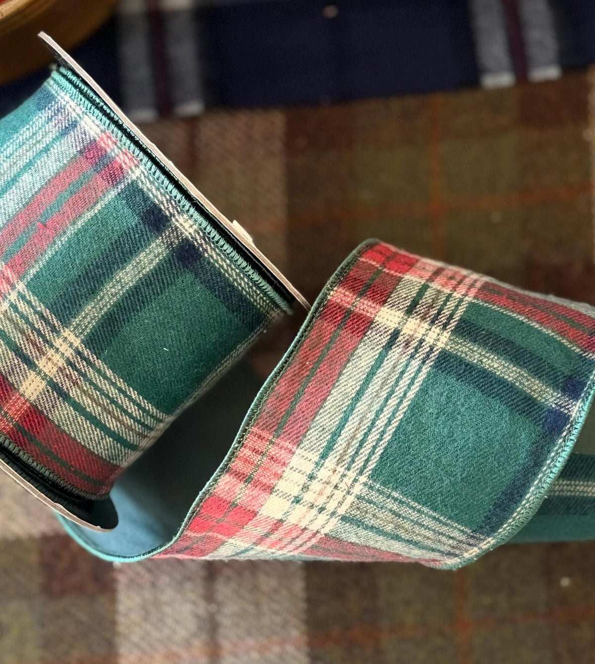 A close-up of a roll of D Stevens Shelton plaid ribbon with a green, red, and cream tartan pattern, shown against a Harris Tweed fabric covered table. 
