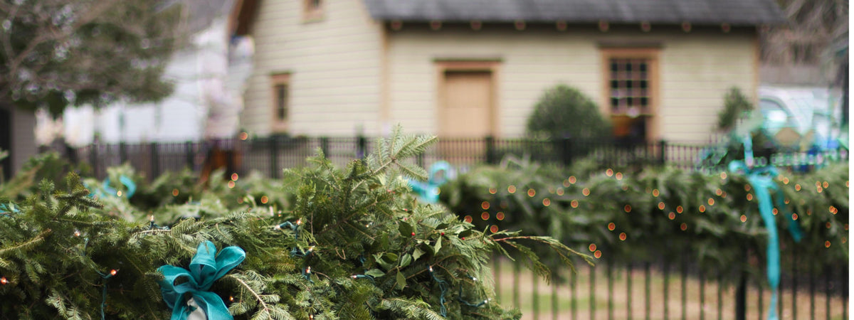 A festive, handmade  green evergreen garland with a bright teal ribbon bow and brass bells in the foreground, with more wreaths and a blurred 1800s carriage house in the background