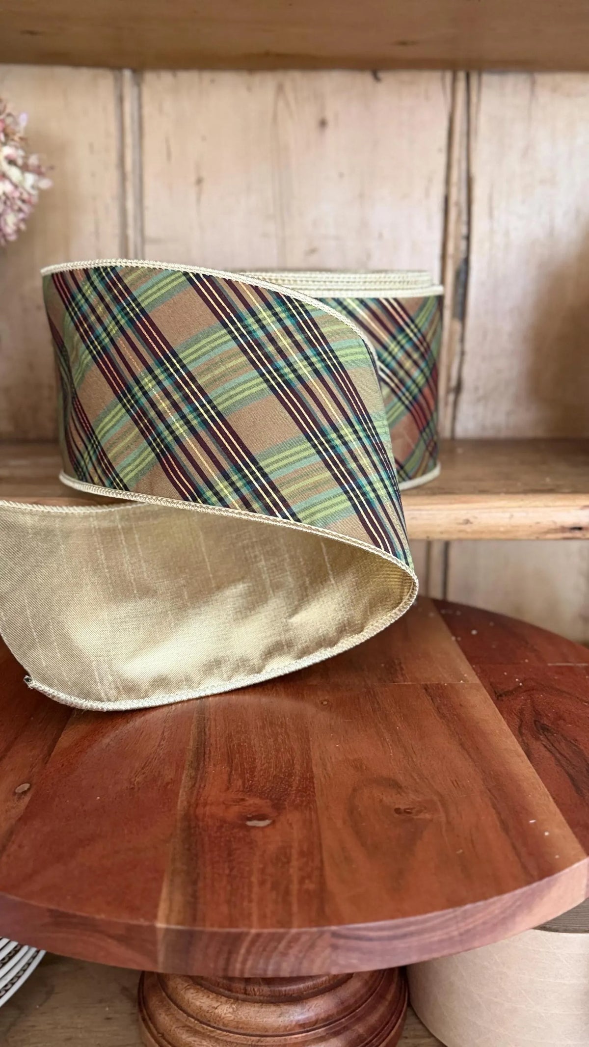A roll of plaid ribbon with a metallic gold satin edge, displayed on a wooden table in front of a wooden antique English pine shelf.