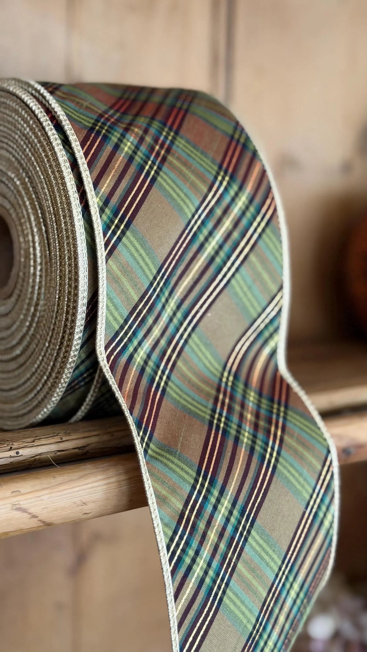 A roll of plaid ribbon draped over a wooden surface, showing a multicolor tartan pattern with green, blue, red, and beige stripes, set against a rustic wooden background.