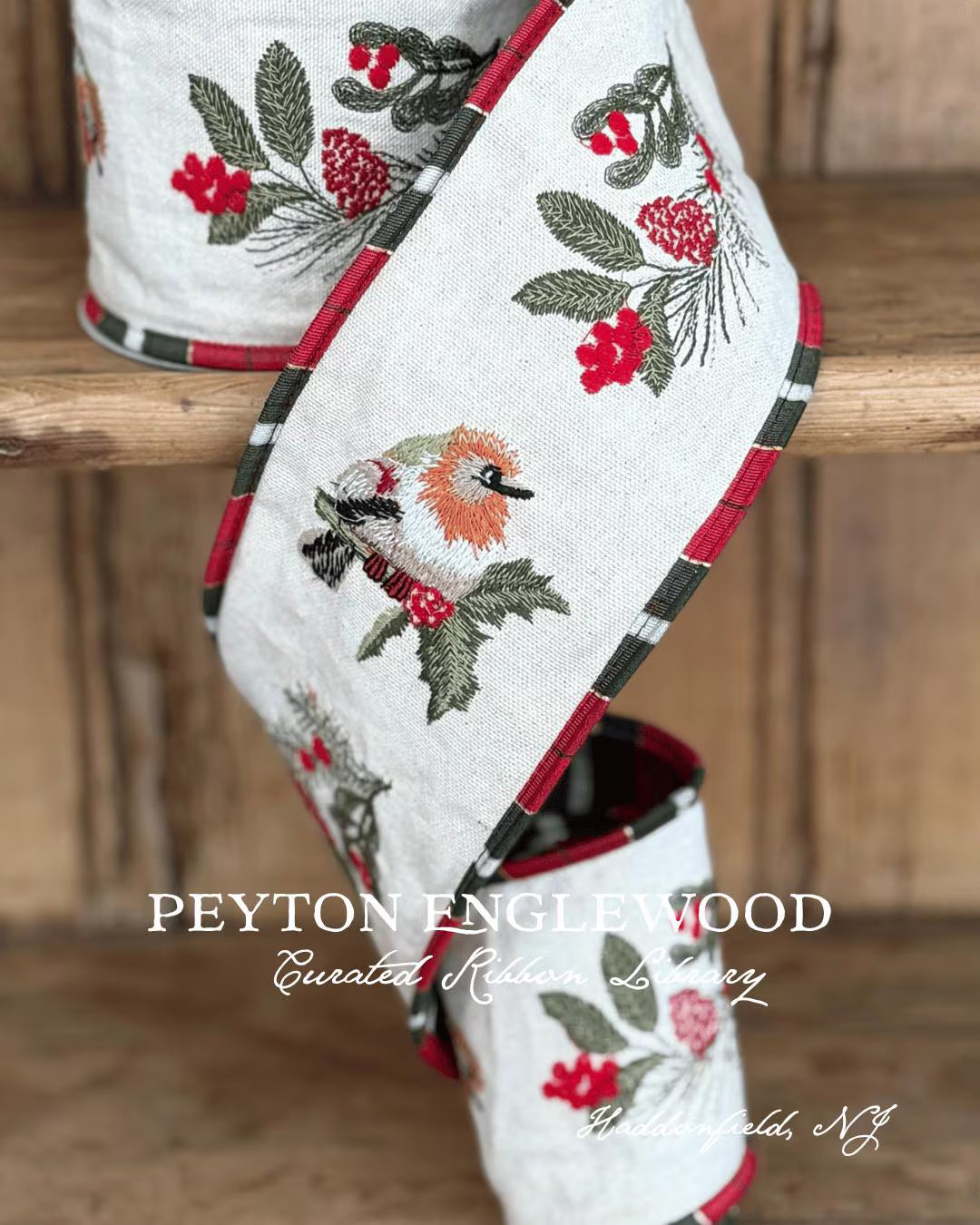 A close-up of a decorative woven textile runner draped over a wooden surface, showing a white base with embroidered birds, red berries, green leaves, and red/black plaid edging. The text 'PEYTON ENGLEWOOD Curated Ribbon Library Haddonfield NJ' is overlaid on the image.