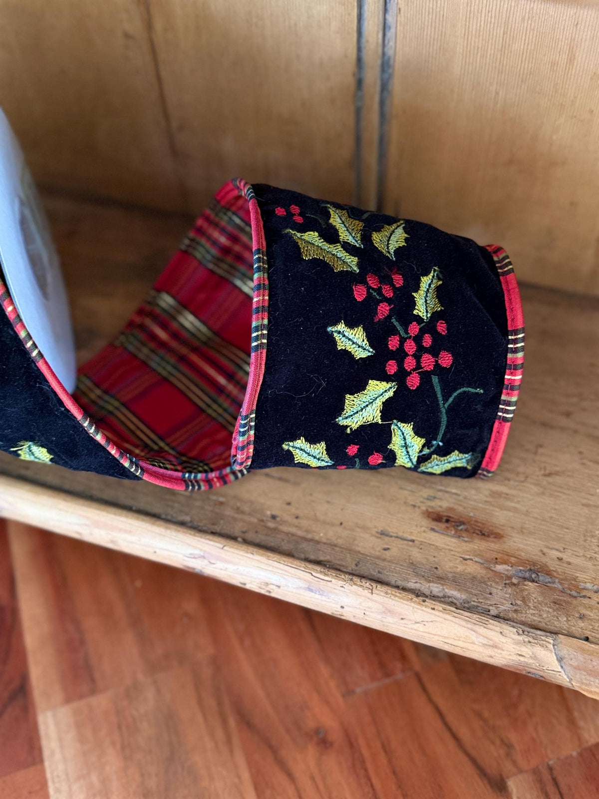 A single black velvet slipper with a red and green plaid lining and a festive embroidered holly and berry pattern on the outer sole, resting on a wooden antique pine surface.